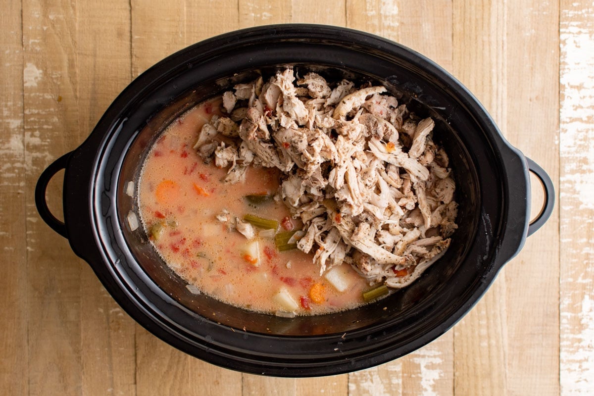 Shredded chicken added to veggies and broth in a slow cooker pot.