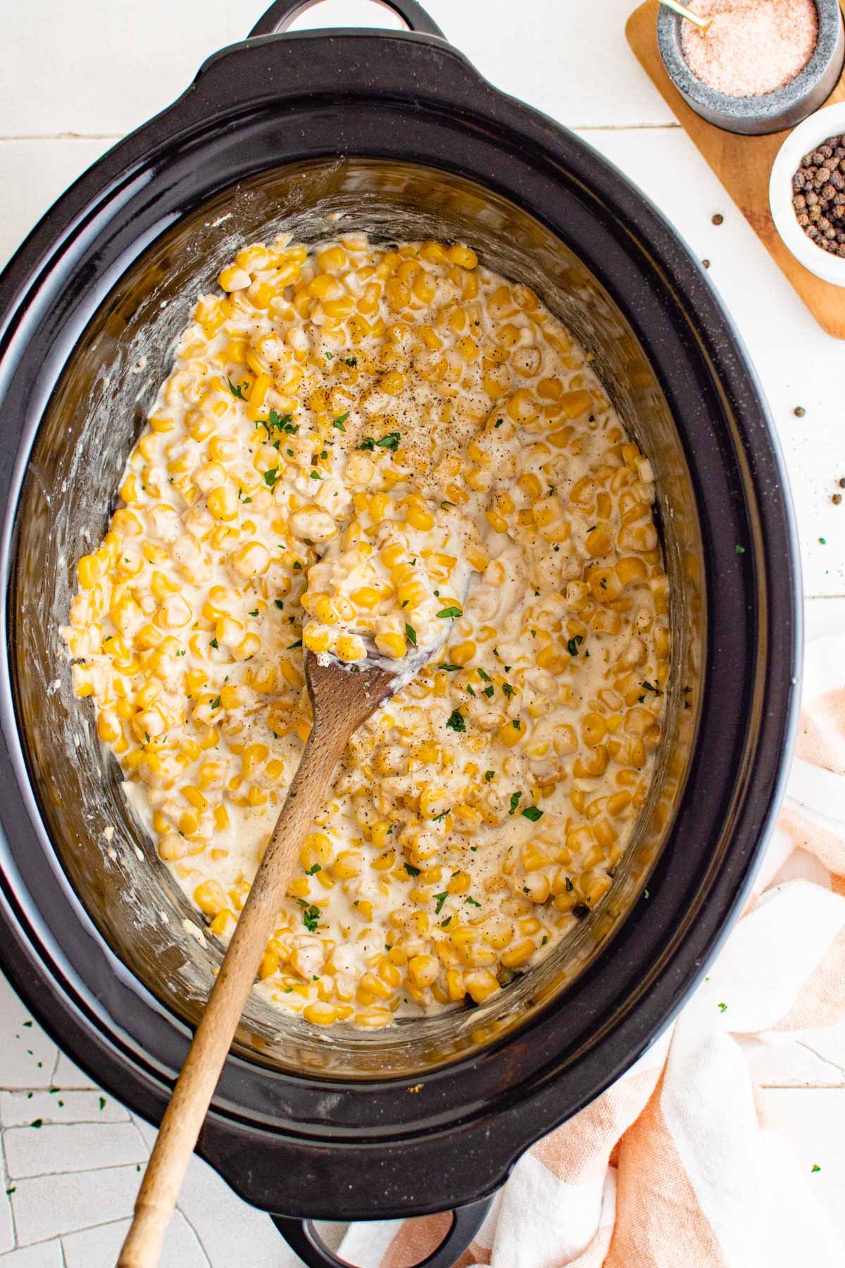 A slow cooker pot, inside it is corn with a creamy sauce and a wooden spoon. 