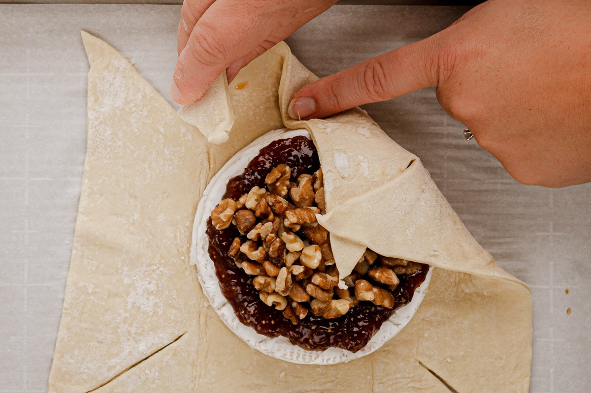 A close up image of puff pastry rolled out on a baking sheet with a wheel of Brie cheese topped with fig jam and walnuts, with the pastry brought up and over the cheese on one side.