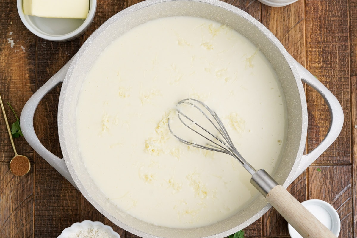 Beshamel sauce in a saucepan with cheeses and a whisk.