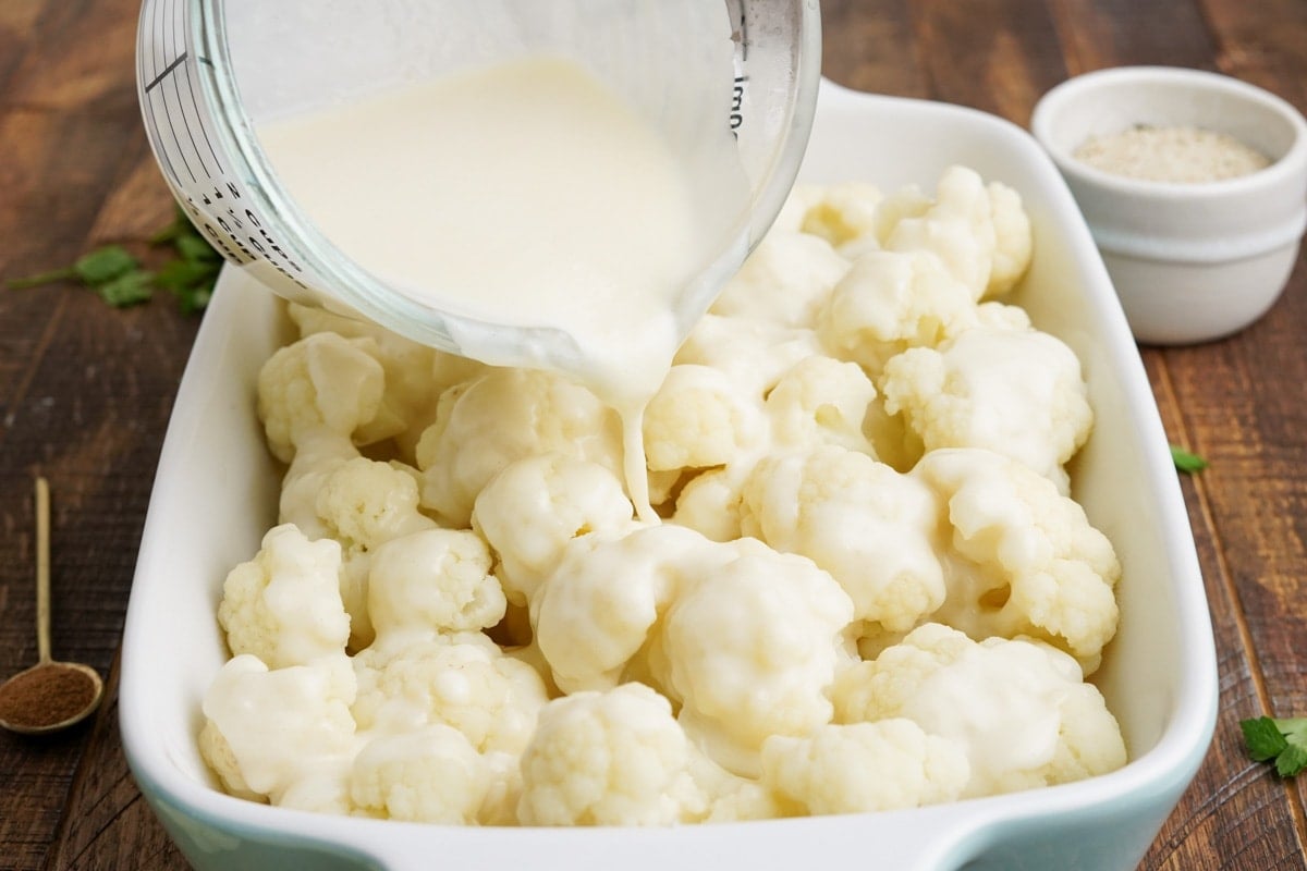 Cheese sauce being poured onto cauliflower in a baking dish.