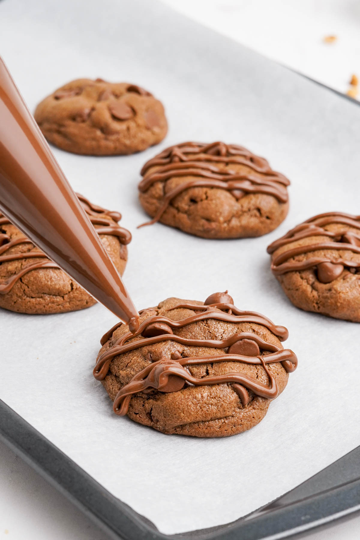 Chocolate cookies on a baking sheet, melted Nutella drizzled from a piping bag.