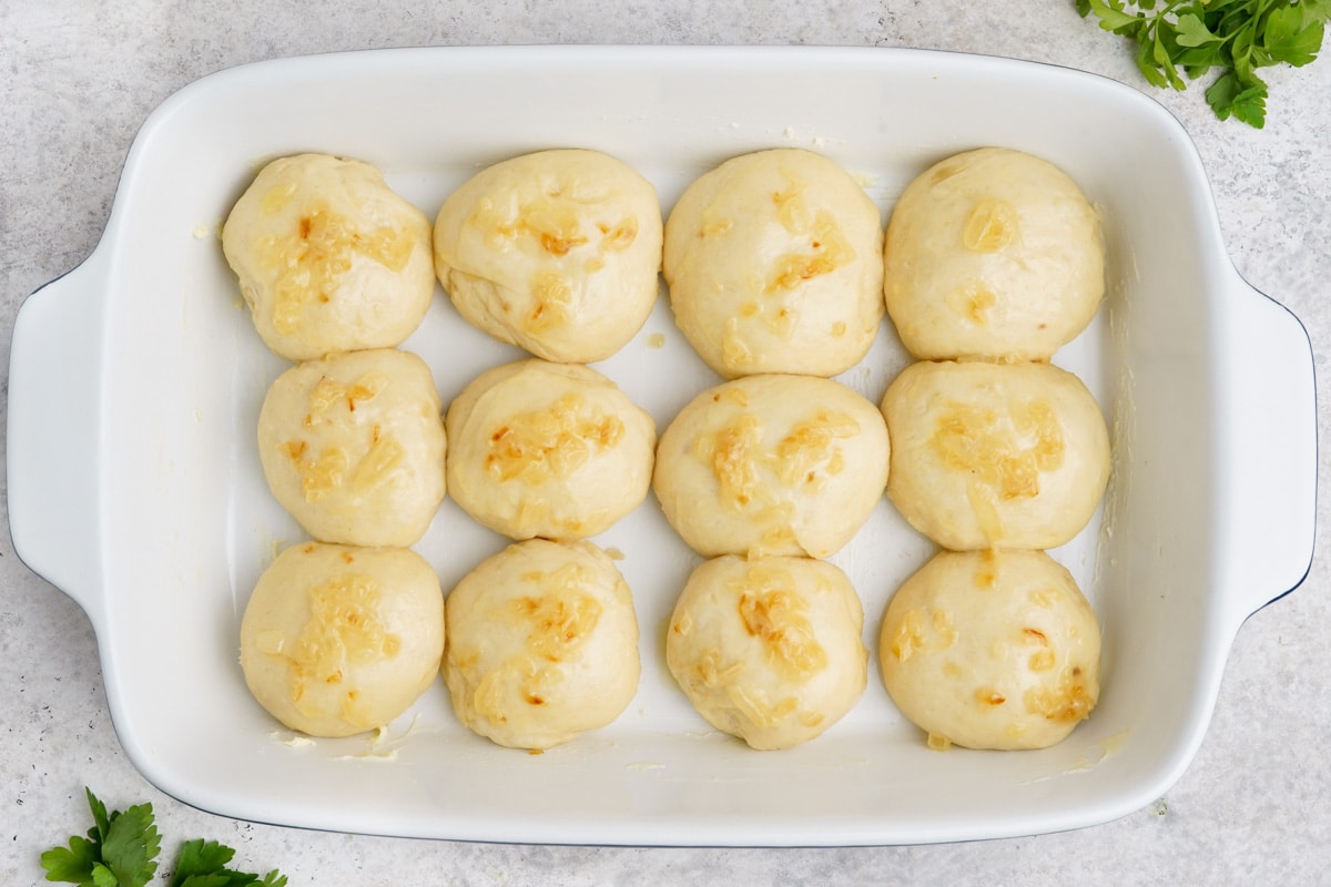 Risen, unbaked rolls with sauteed onions on top, in a white baking dish.