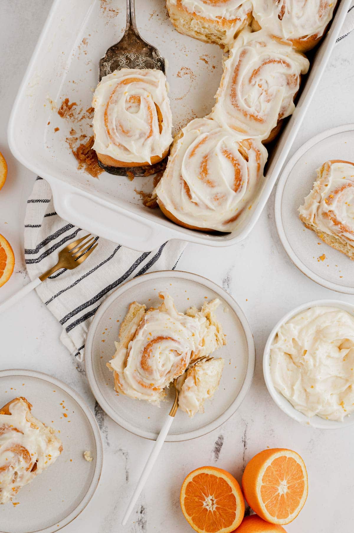 Overhead shot of orange rolls in a baking dish and on plates, sliced oranges with a black and white striped napkin.