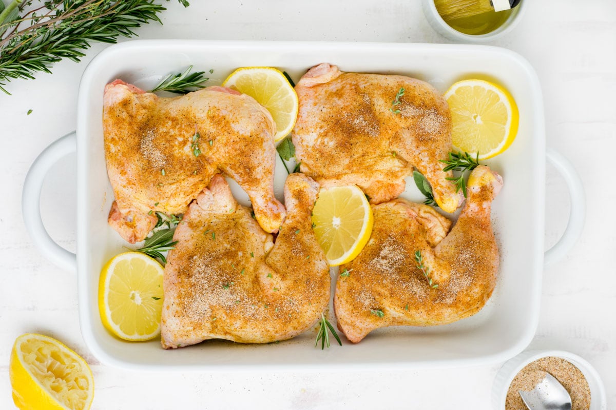 Chicken leg quarters with seasoning and lemon slices on a cutting board.
