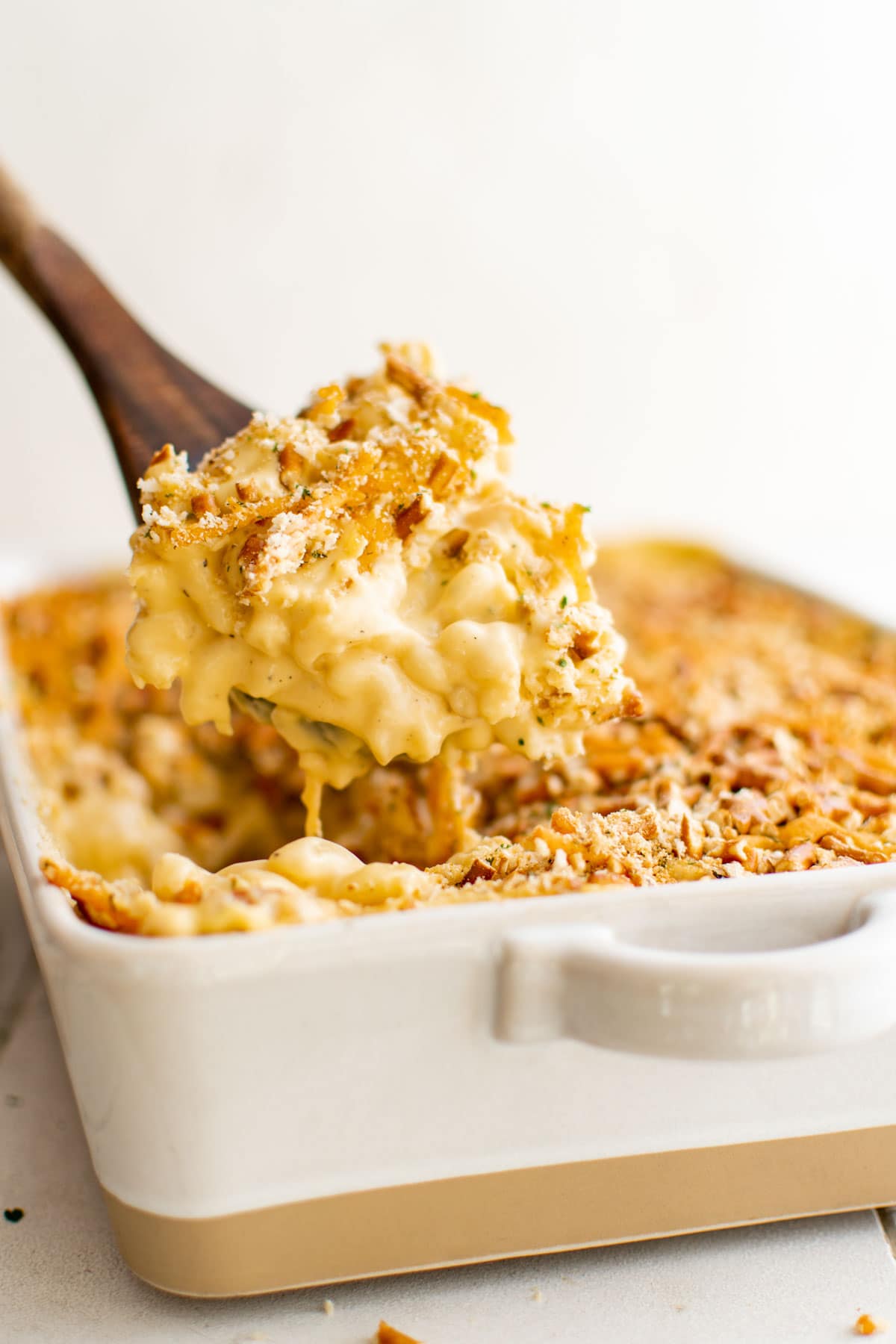 Baked macaroni and cheese in a white ceramic dish with a pretzel crust and a wooden spoon.