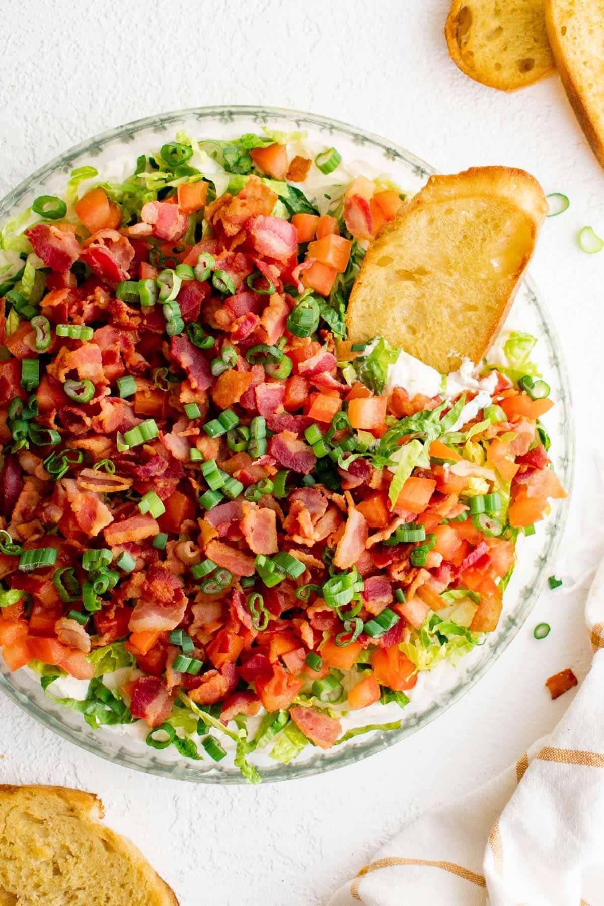 Clear glass pie plate with creamy base, lettuce, tomatoes, bacon and green onions.