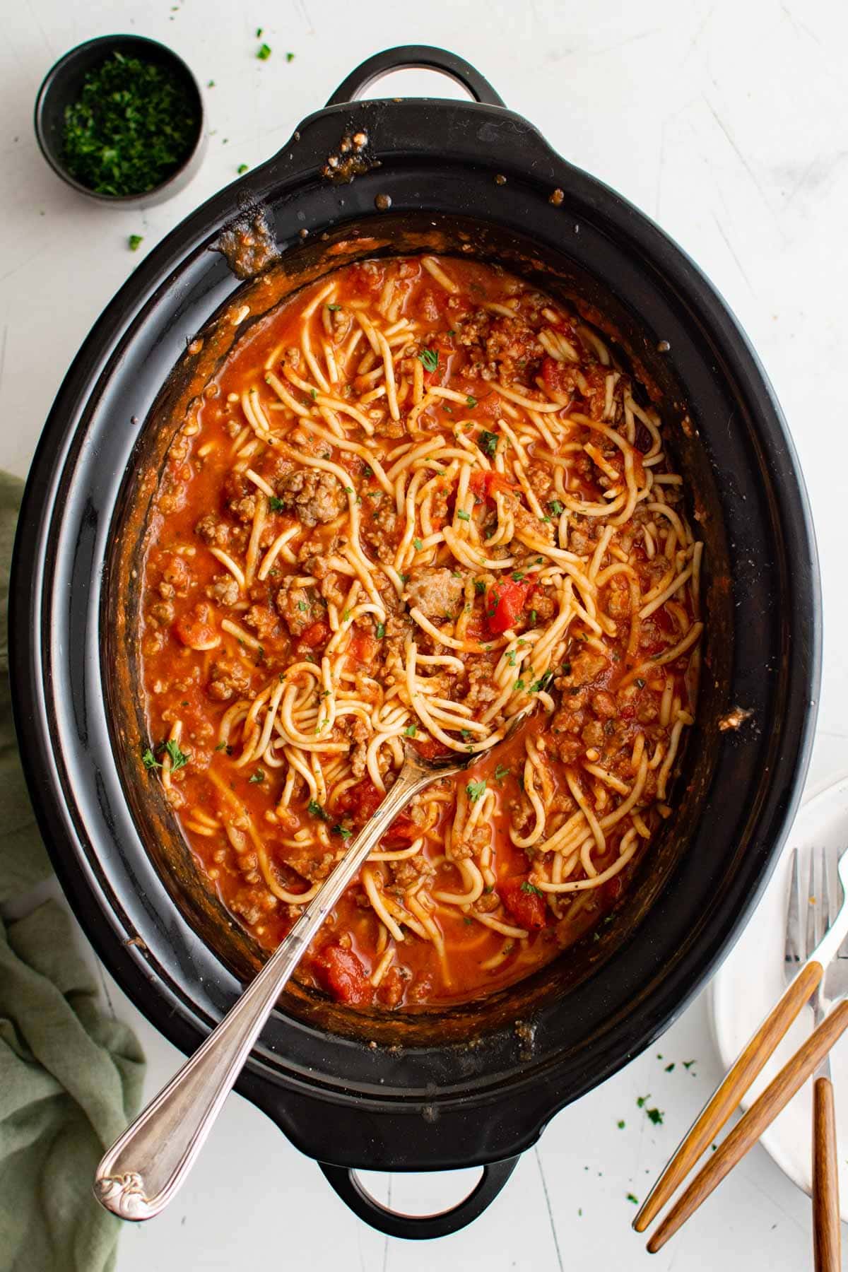 Slow cooker with spaghetti noodles and sauce and a large serving spoon.