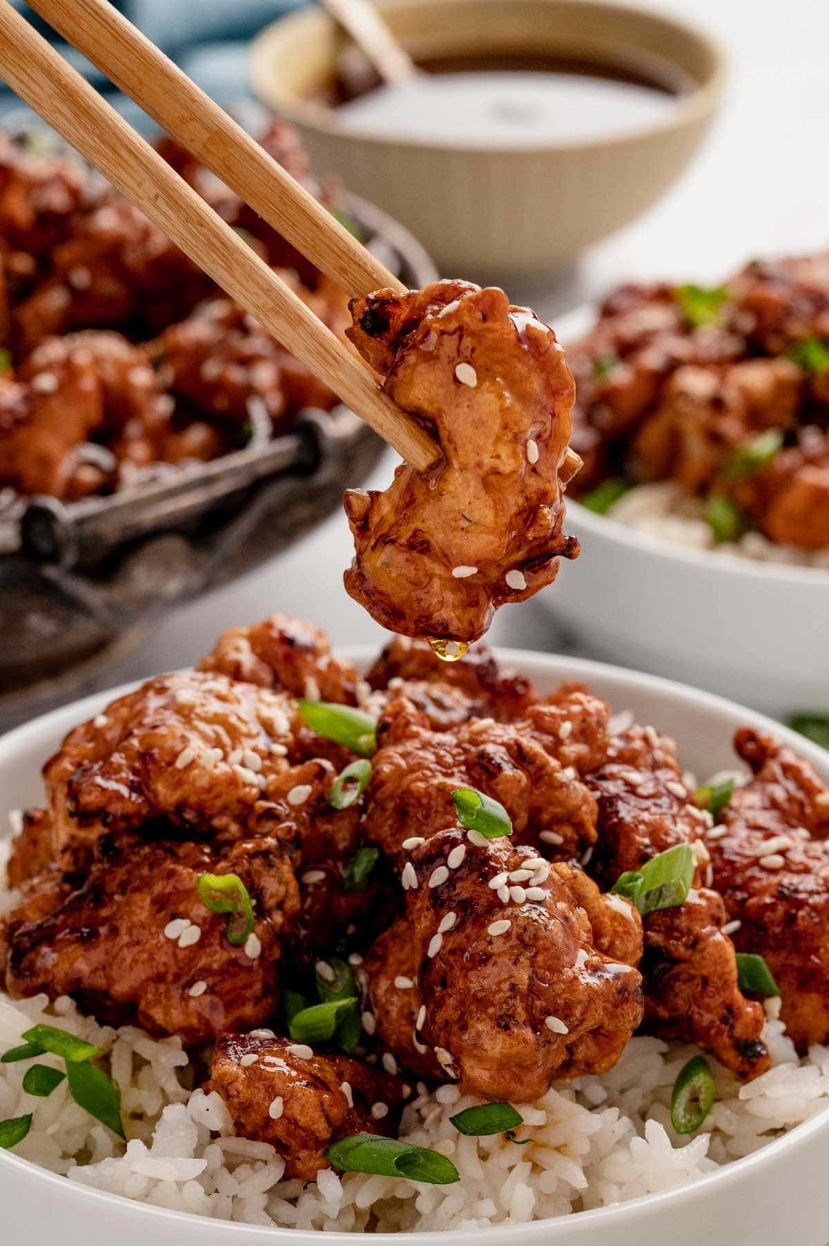 Piece of crispy honey chicken held by chopsticks over a bowl of the same.