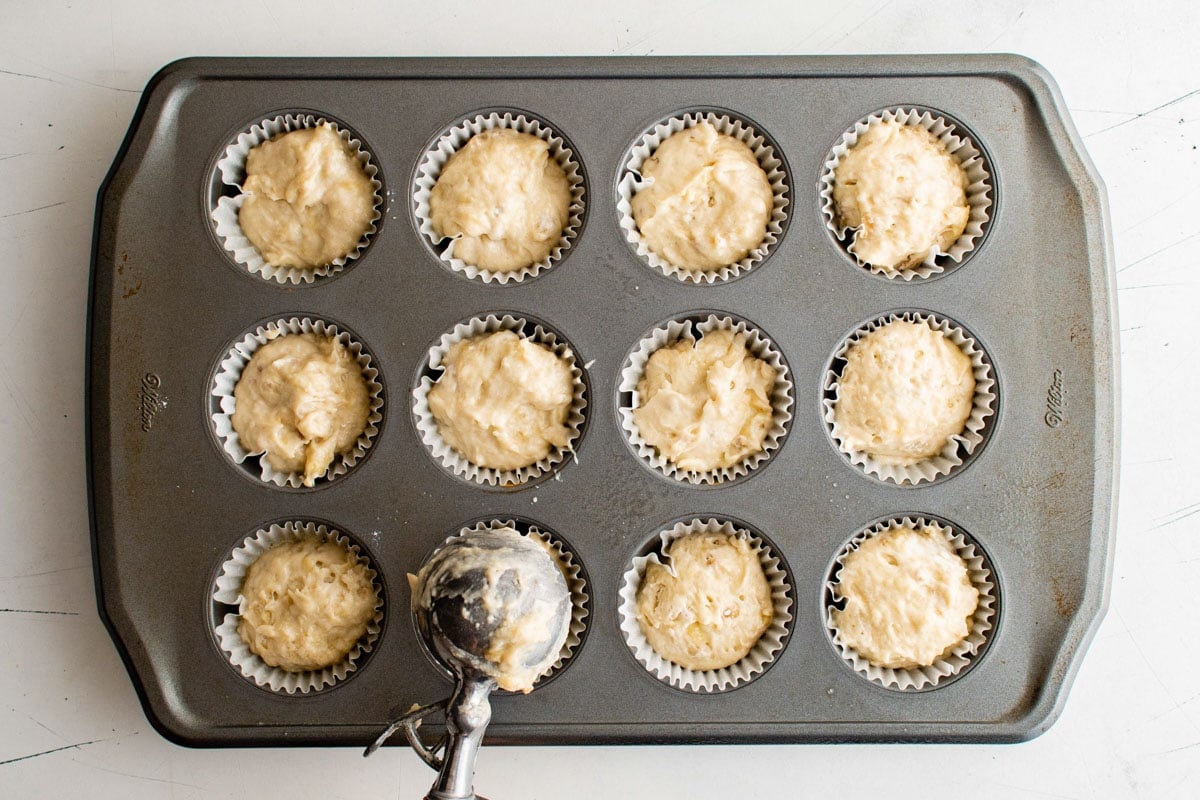 Muffin pan with banana muffin batter and an ice cream scoop for portions.