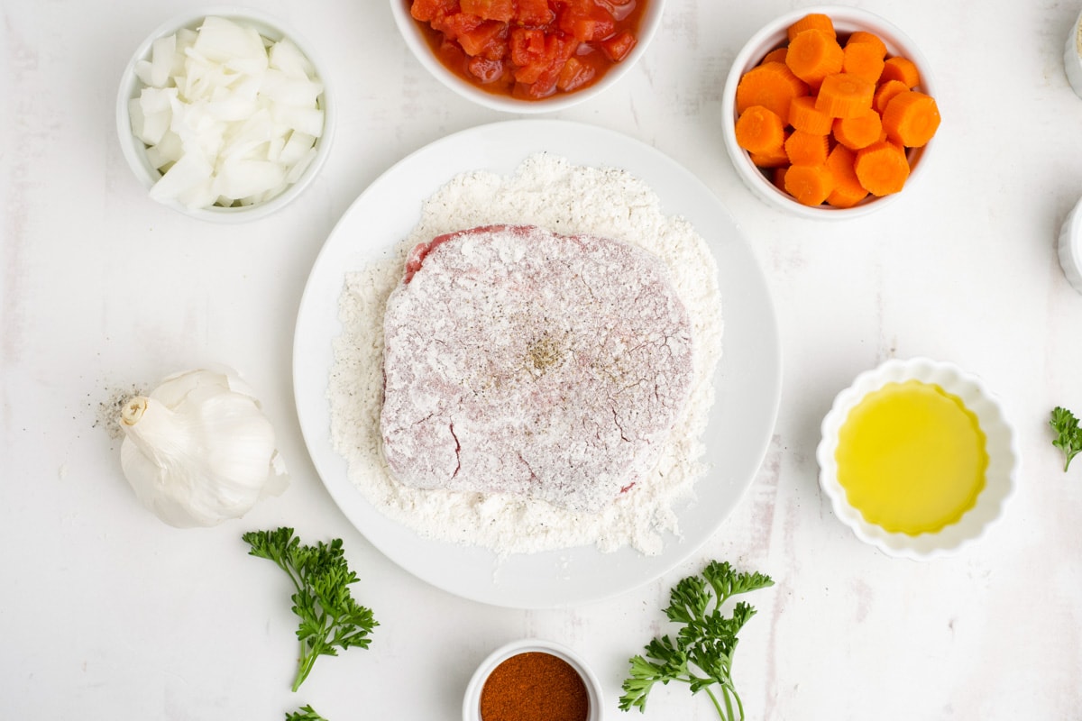 Thin steak coated in flour and surrounded by small dishes of carrots, tomatoes, oil and onions., 