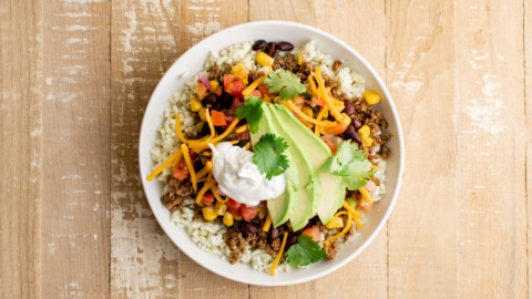 Taco bowl with rice, ground beef taco meat, beans, corn, cheese, tomatoes, sour cream and avocado.