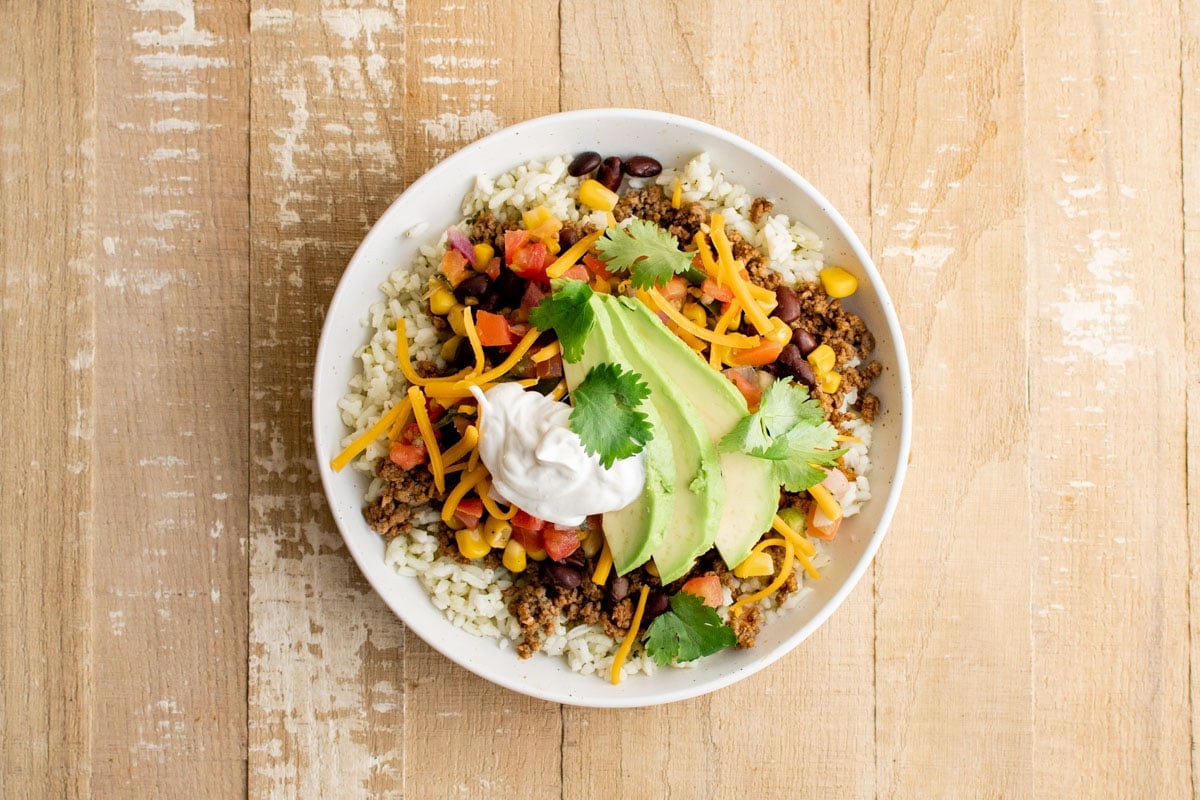 Taco bowl with rice, ground beef taco meat, beans, corn, cheese, tomatoes, sour cream and avocado.