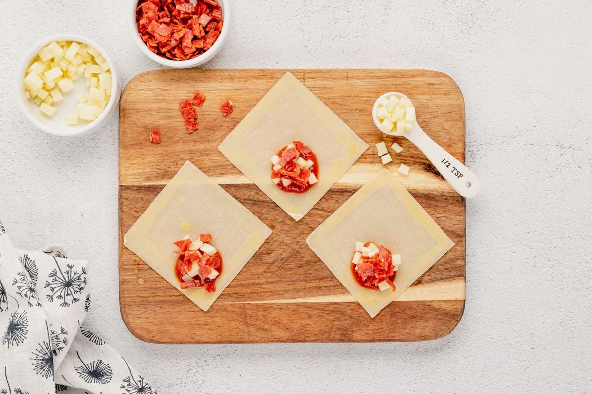 eggroll wrappers on a cutting board with sauce, pepperoni and mozzarella. 