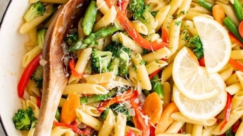 Penne pasta and vegetables in a large skillet with lemon slices and a wooden spoon.