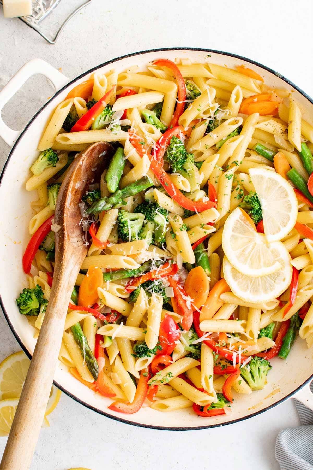 Penne pasta and vegetables in a large skillet with lemon slices and a wooden spoon.