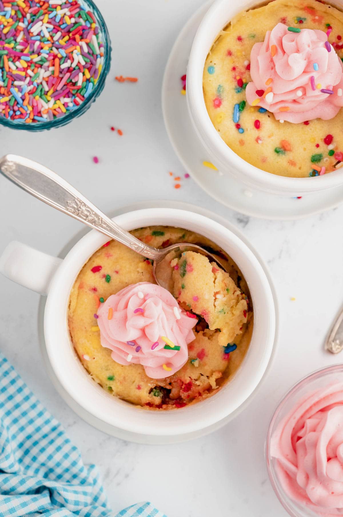 Mug cakes, vanilla with sprinkles and pink frosting, in white mugs.