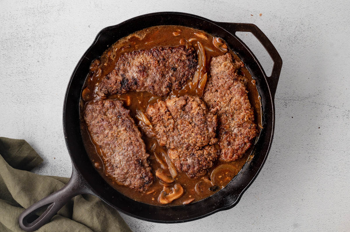 Sauteed cub steaks set into gravy with mushrooms and onions in a skillet.