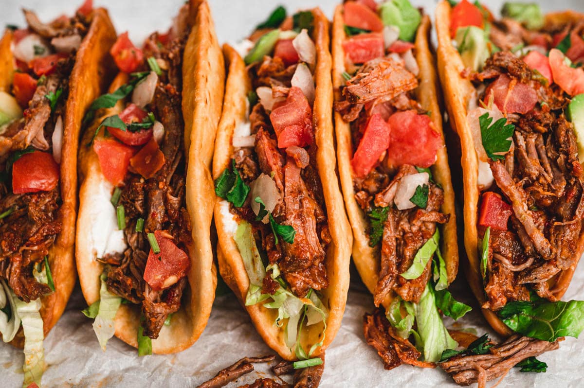 Crispy taco shells lined up and filled with shredded beef topped with tomatoes.