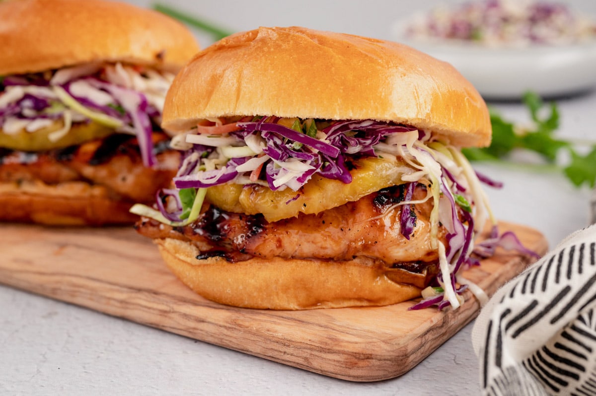 teriyaki chicken sandwich with coleslaw and pineapple on a wooden cutting board.