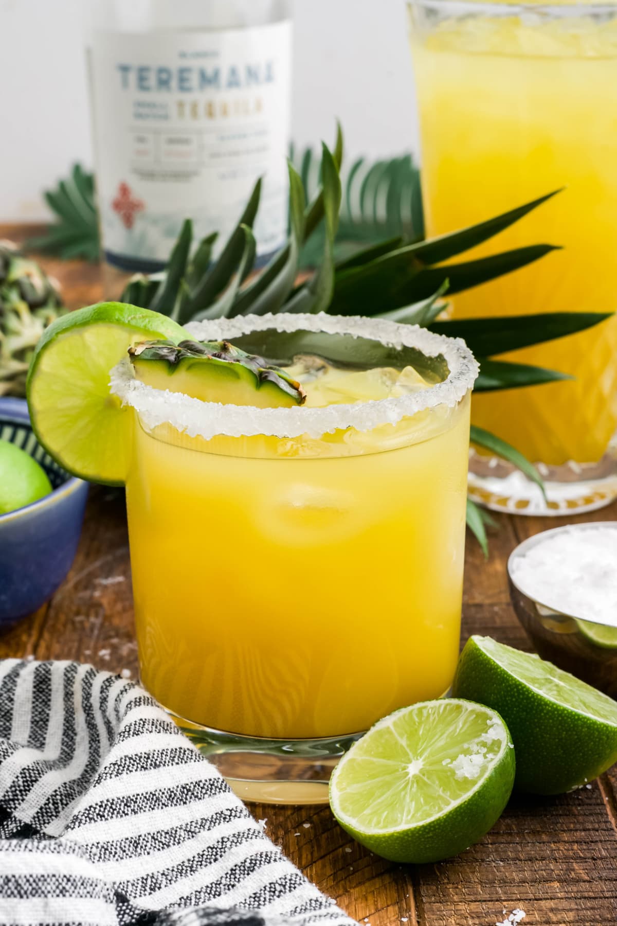 Cocktail glass with pineapple margarita, limes and pineapple, and a bottle of tequila in the background.