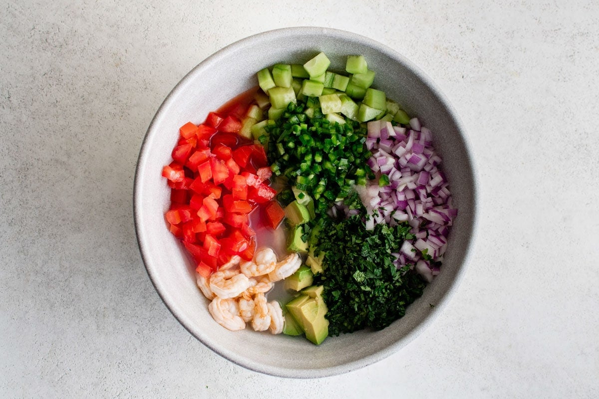 Diced tomatoes, cucumber, cilantro, shrimp, onion and avocado in a large white bowl.