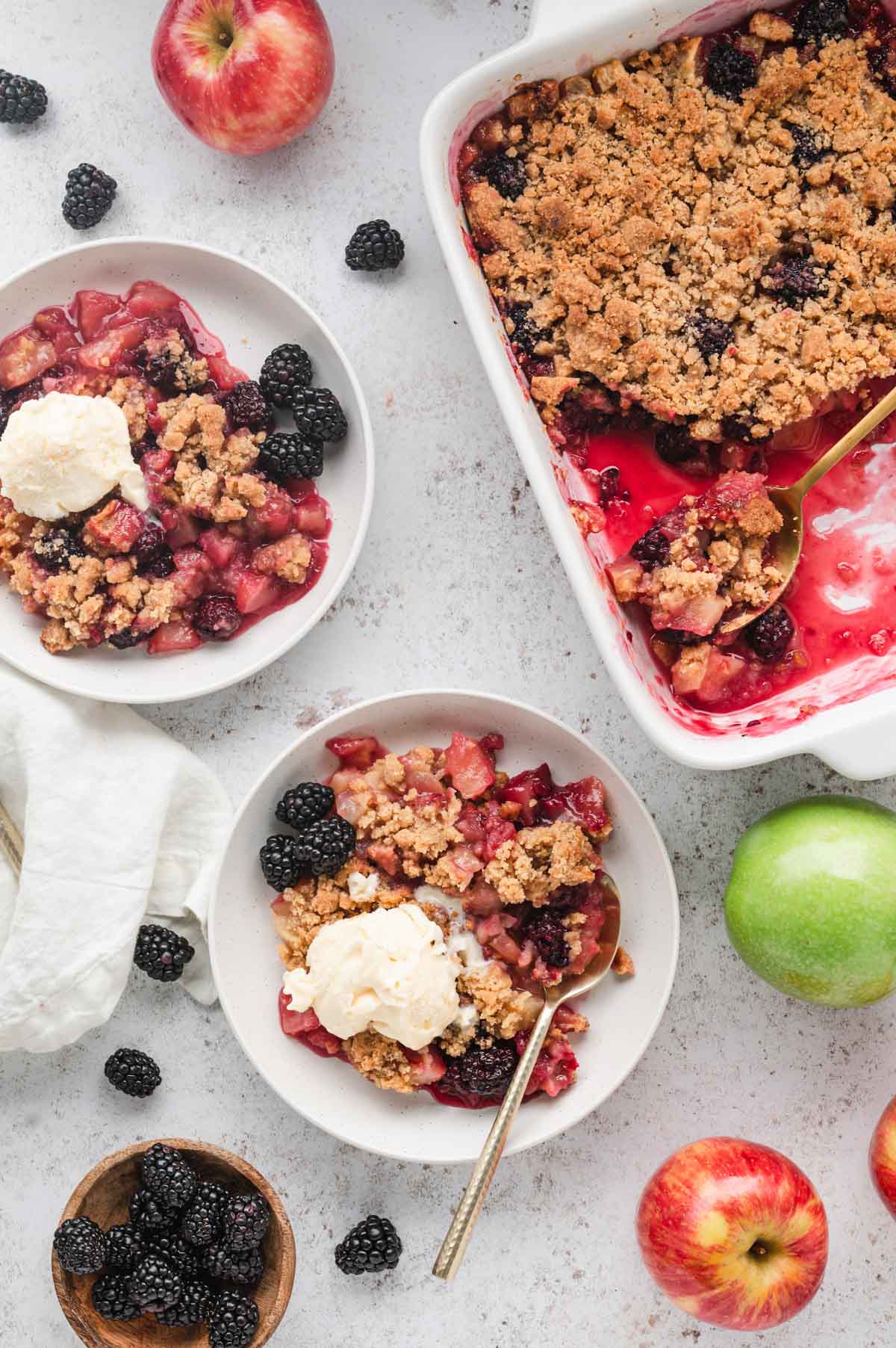 Apple and blackberry crumble with vanilla ice cream in white bowls with spoons.