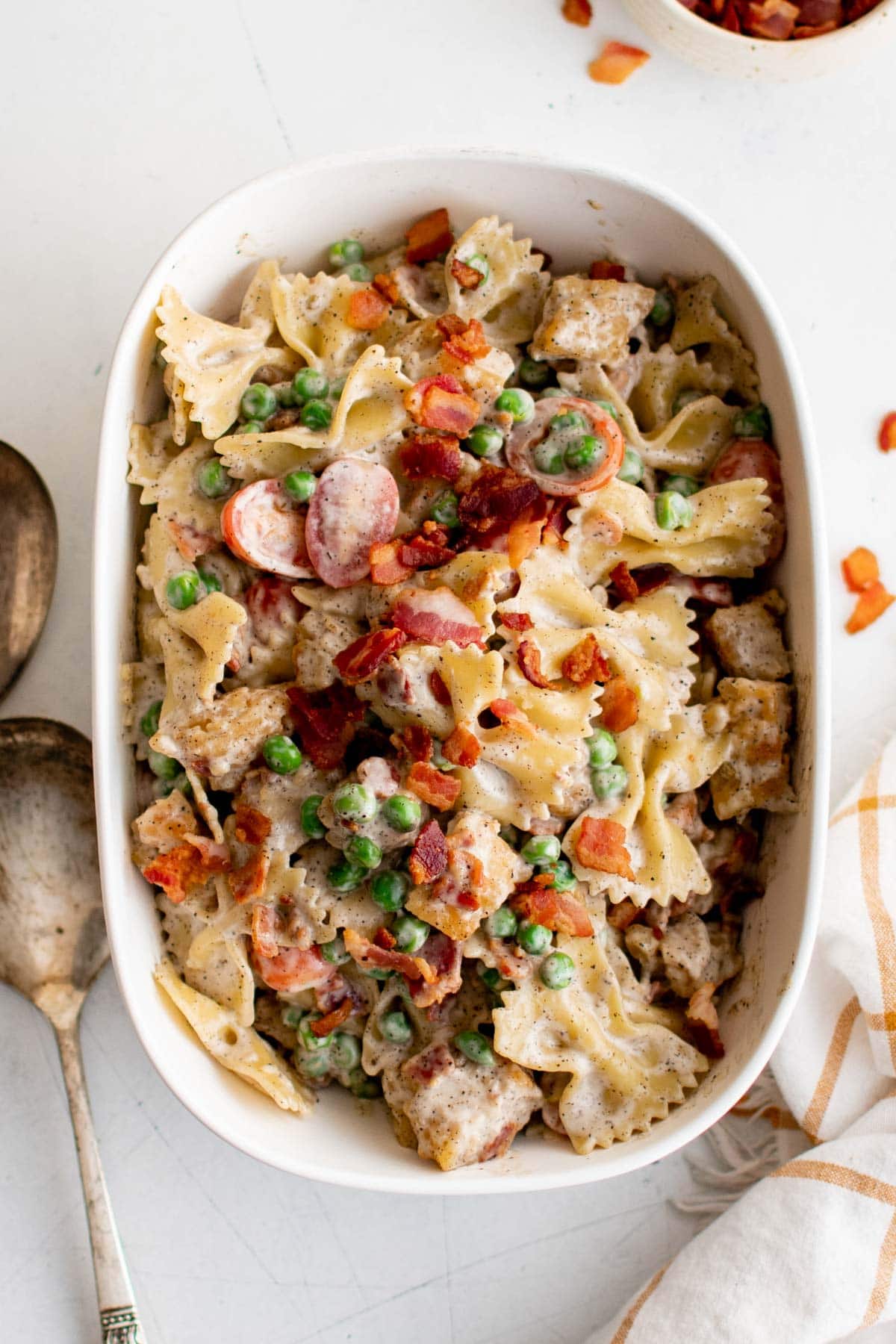 Pasta, peas, bacon and tomatoes and chicken mixed with a dressing and presented in a white serving dish.