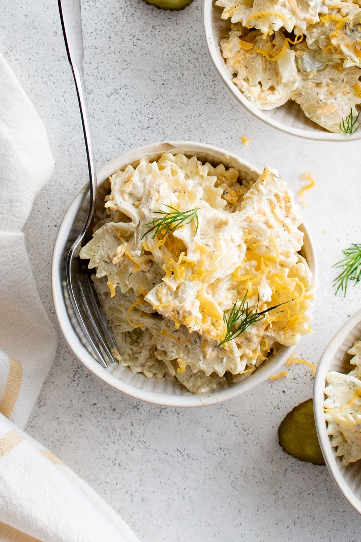 Small white bowl with pasta salad with dill pickles and shredded cheddar and a fork.