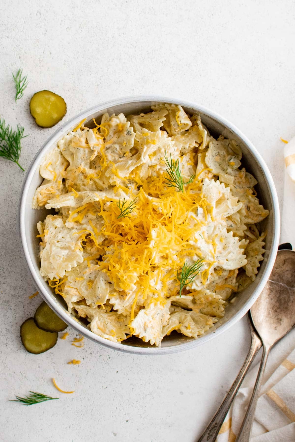 White serving bowl, bowtie pasta with creamy dressing, fresh dill and shredded cheddar cheese.