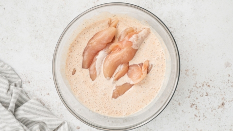 Chicken tenders in a seasoned buttermilk in a glass bowl.