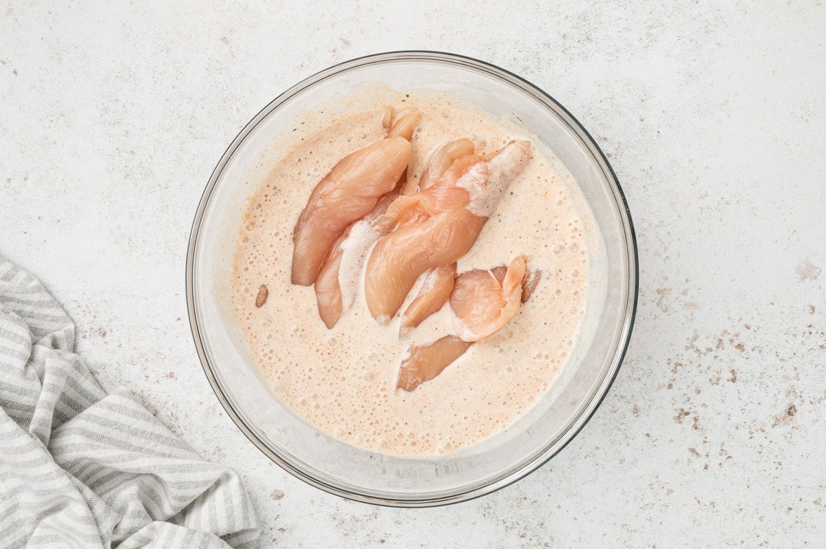 Chicken tenders in a seasoned buttermilk in a glass bowl.