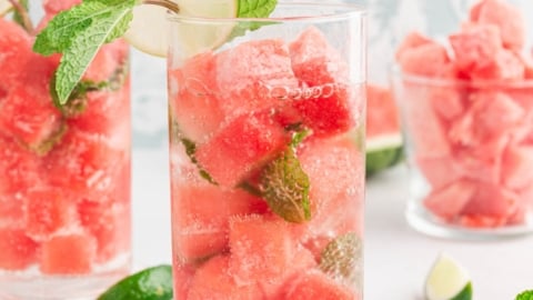 Clear beverage in a tall glass with watermelon ice cubes, mint and limes.