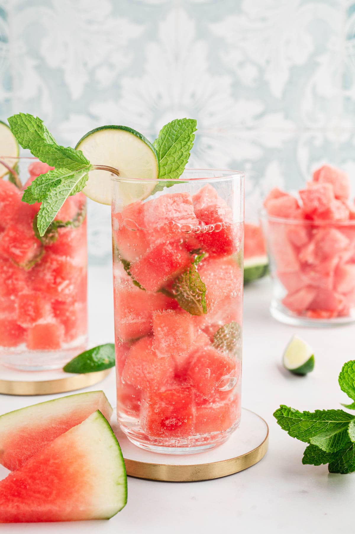 Clear beverage in a tall glass with watermelon ice cubes, mint and limes.