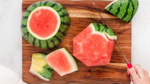 Watermelon on a cutting board, with a serrated knife cutting the rind off.