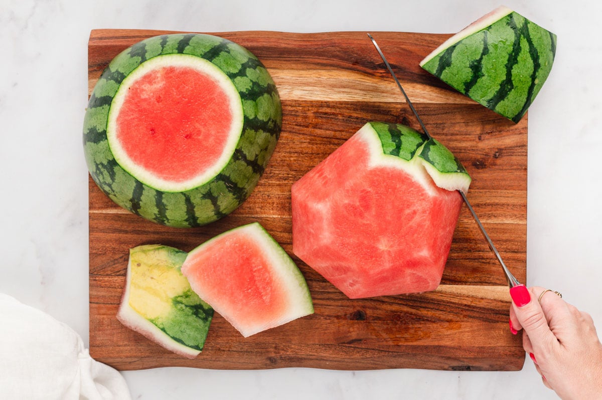 Watermelon on a cutting board, with a serrated knife cutting the rind off.