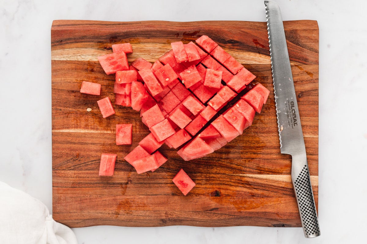 Watermelon cubes and a serrated knife on a cutting board.