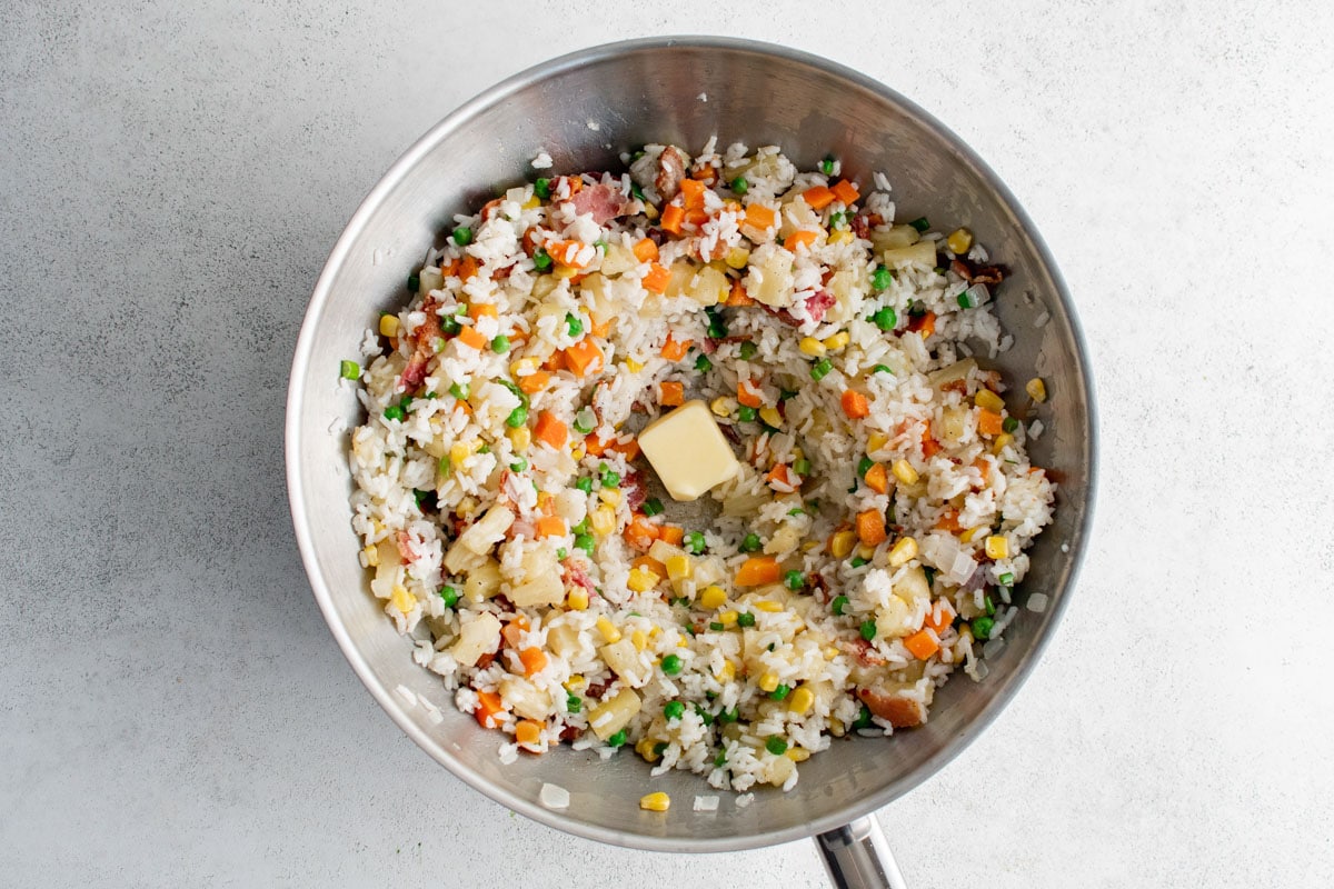 Rice, veggies, pineapple and bacon combined in a wok, with a pat of butter in the center.