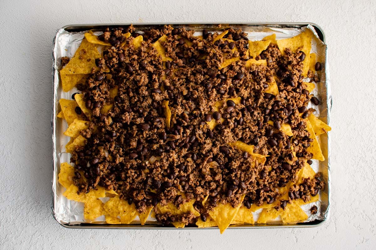 Ground beef on top of tortilla chips on a foil covered sheet pan.