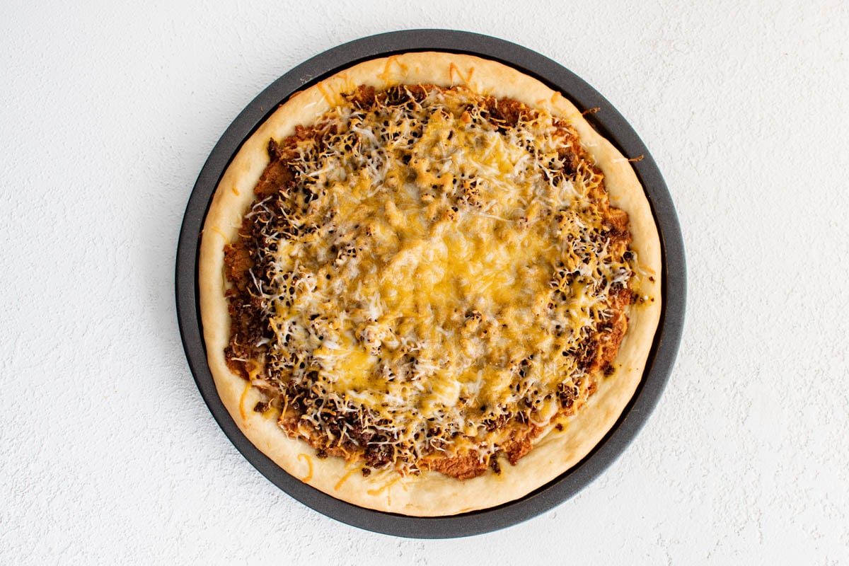 Baked pizza crust with refried beans, taco meat and cheese.