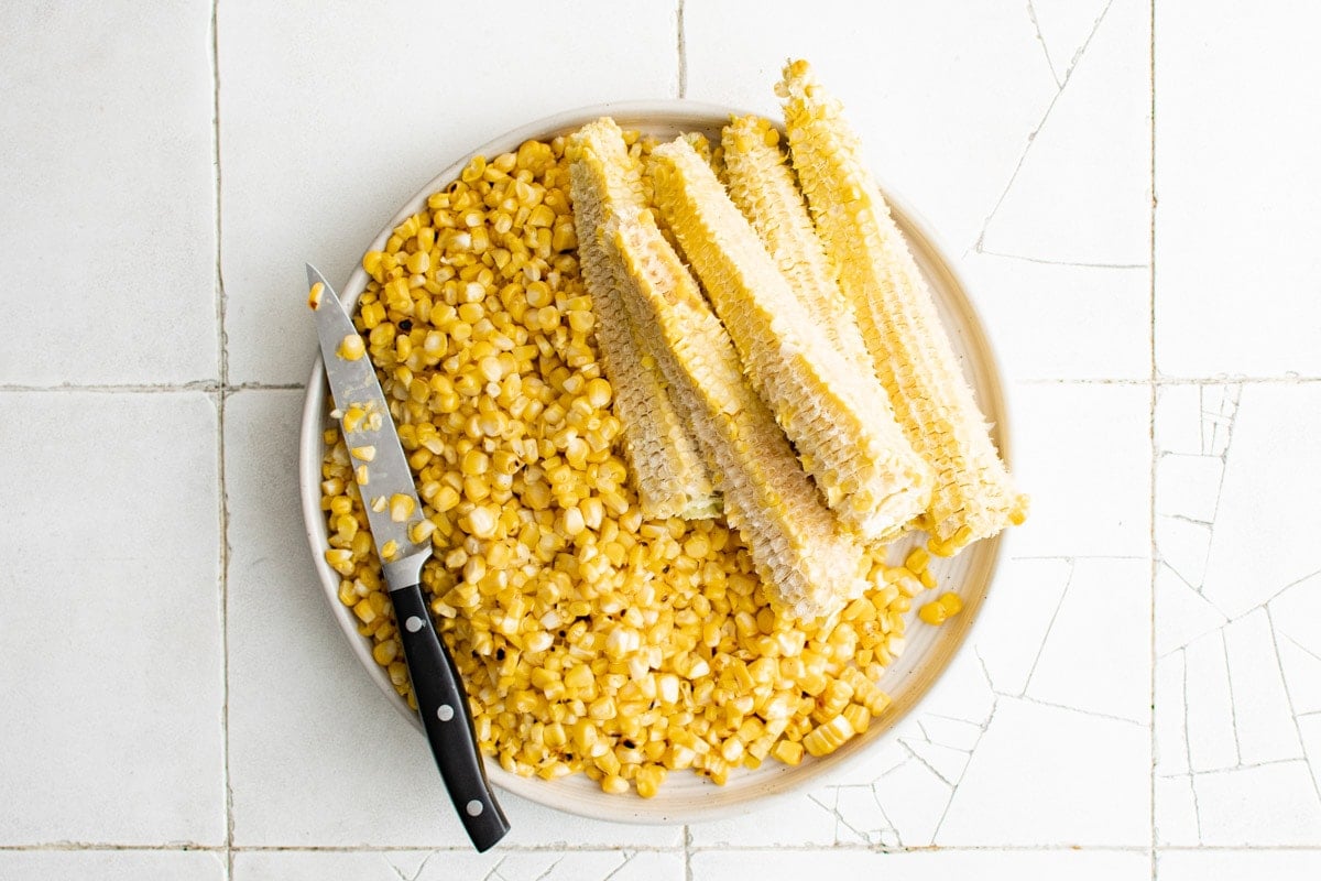 Empty corn cobs, a bowl of corn cut off the cobs, and a knife.