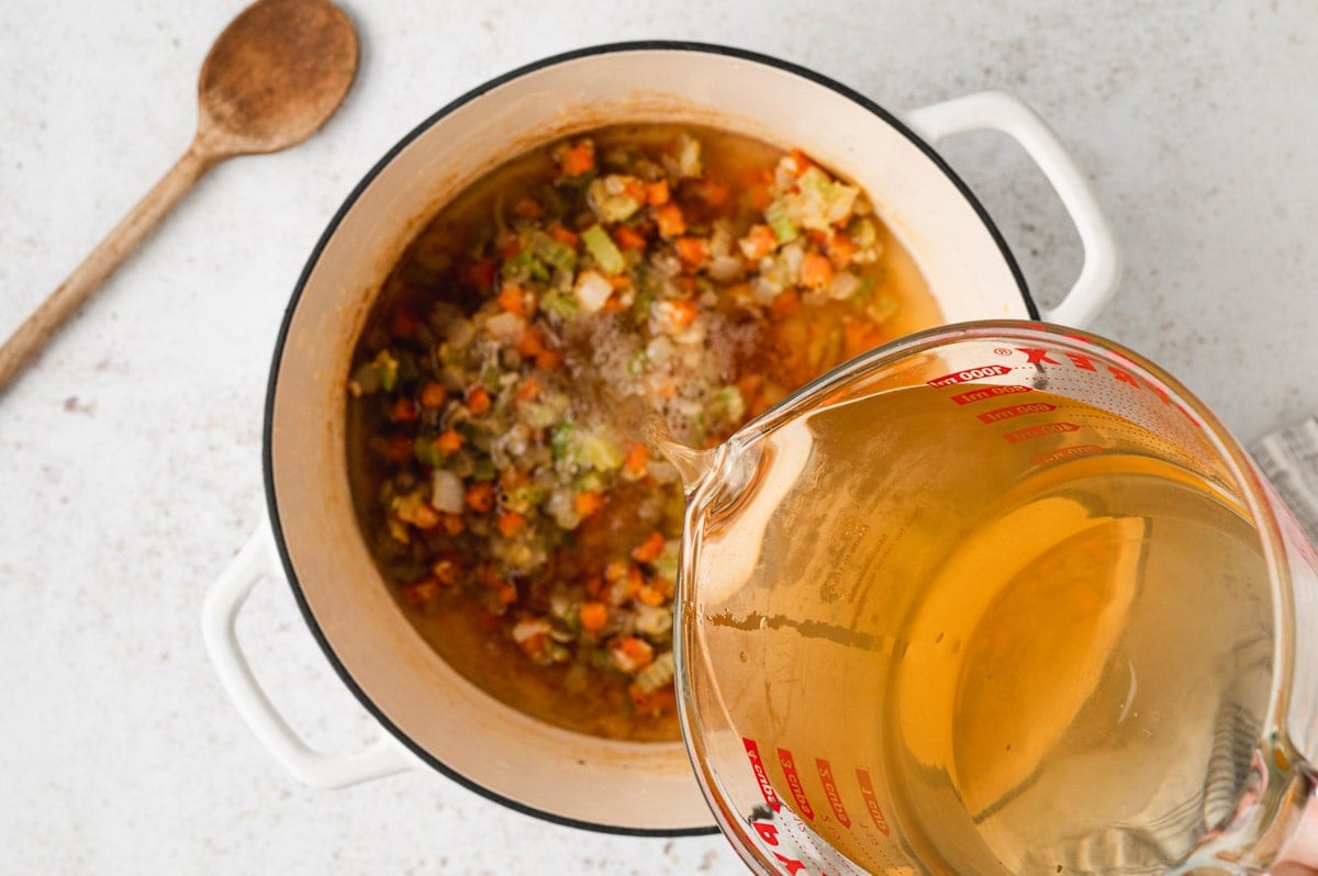 Chicken broth being poured into a soup pot with carrots, celery and onions.