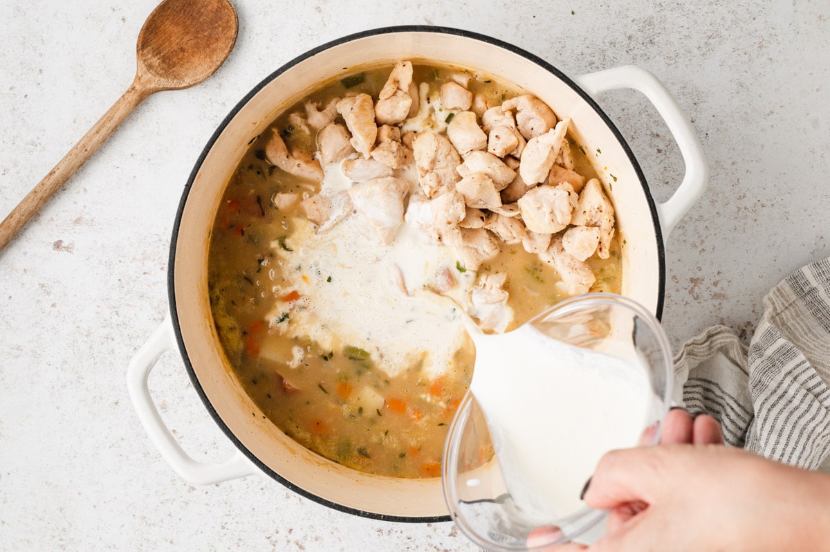 Cream being poured into a large pot with chicken, vegetables and broth.