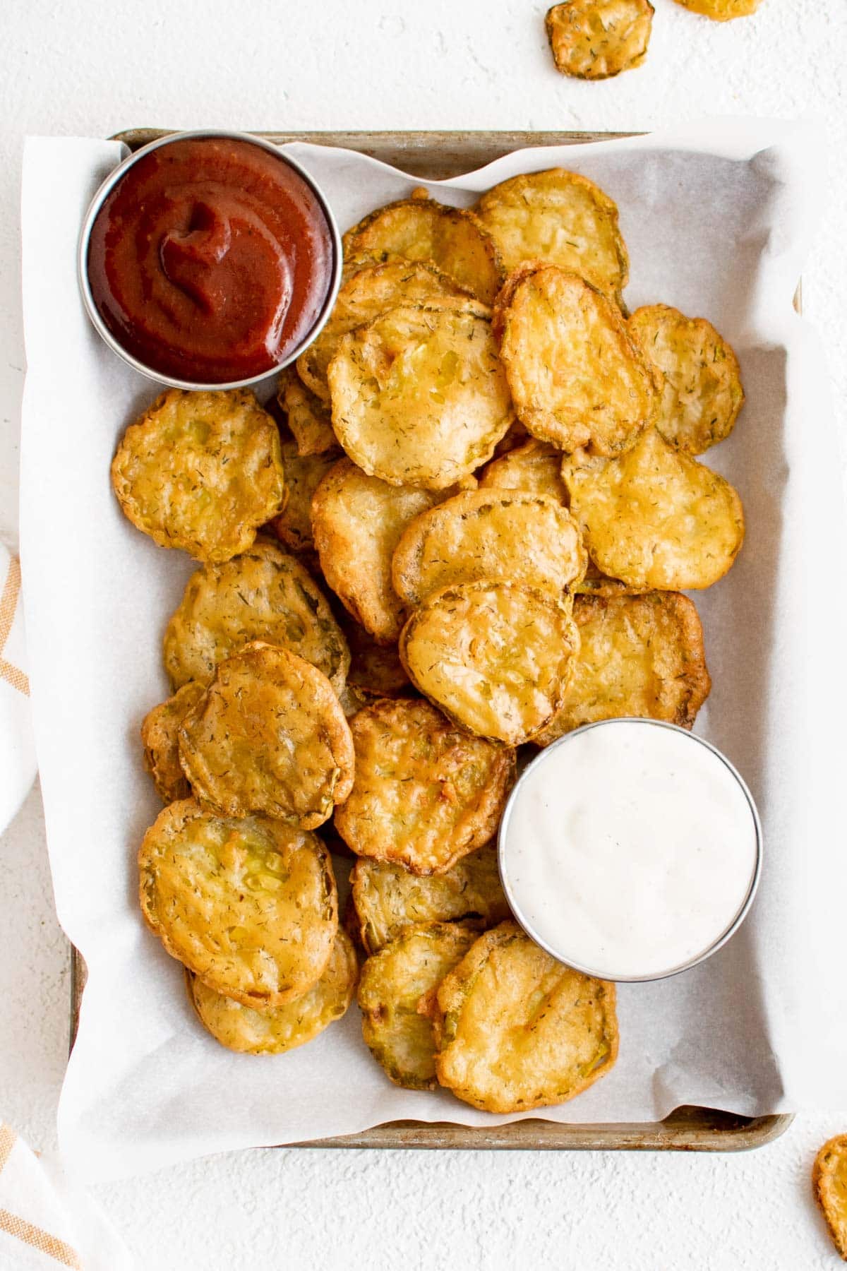 Fried dill pickle chips on a small tray with parchment paper and a small cup of ranch and a small cup of bbq sauce.