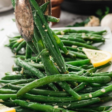 Green beans stacked on a platter and lifted with silver metal serving spoons.