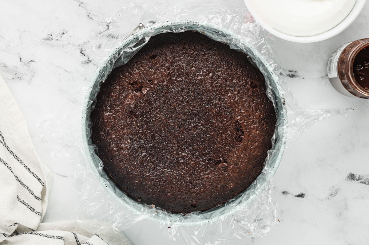 Baked chocolate cake in a round pan with plastic wrap.