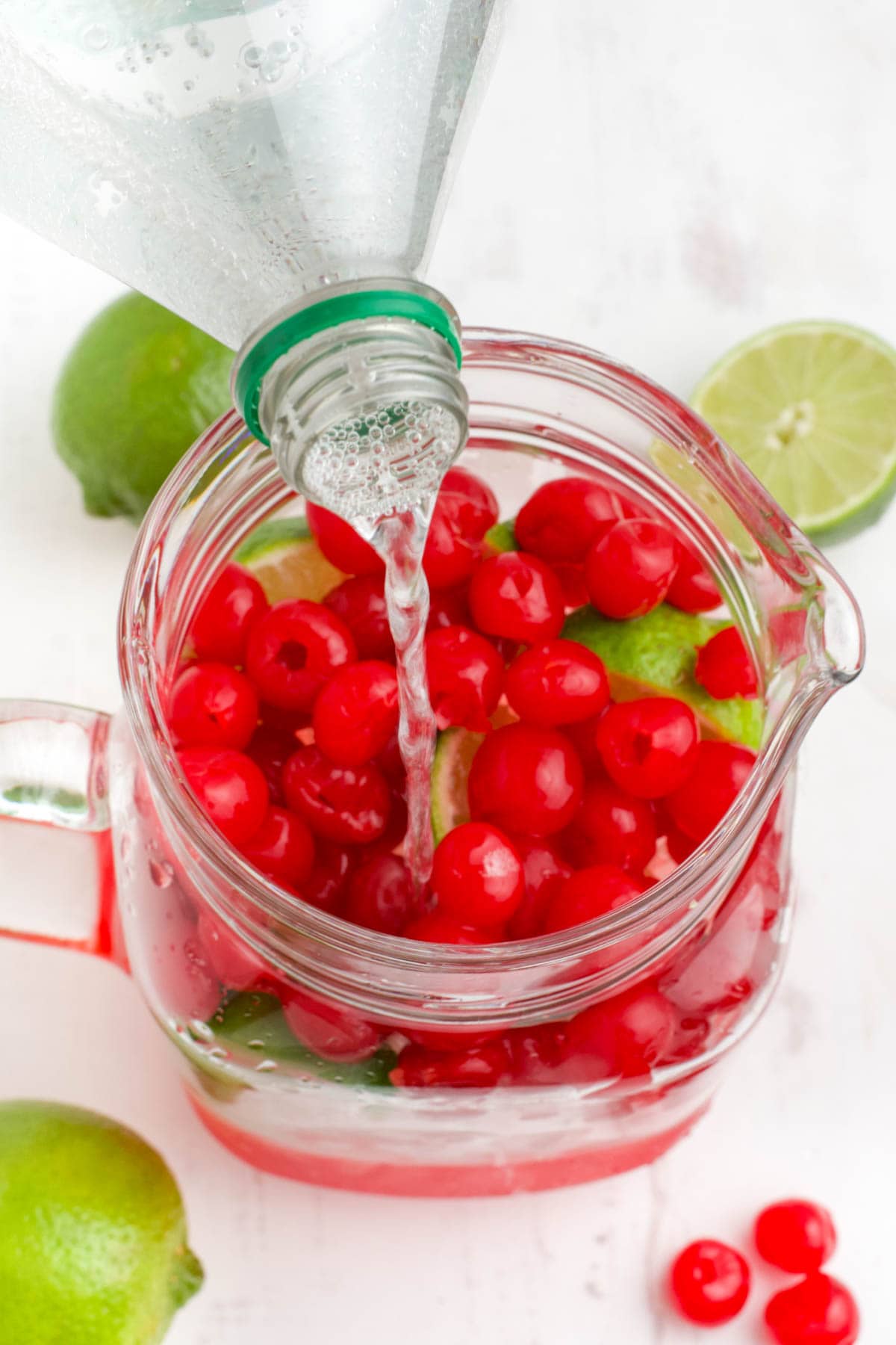 Pouring Sprite from a bottle into a large pitcher of cherries and limes.