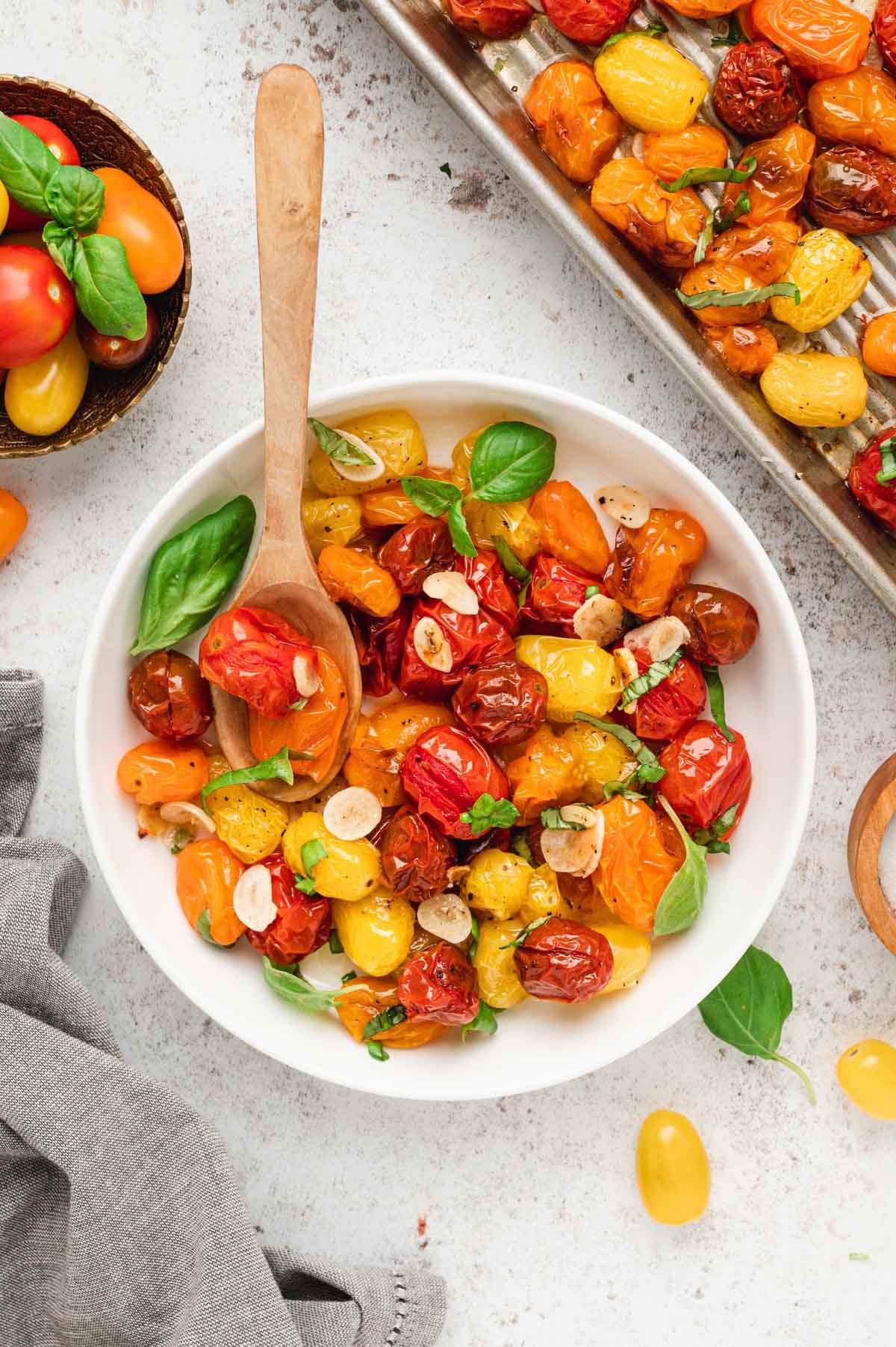 Roasted cherry tomatoes in a white bowl with fresh basil leaves and a wooden spoon.
