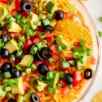 Square image of round dish with shredded lettuce, cheese, olives, tomatoes and green onions.