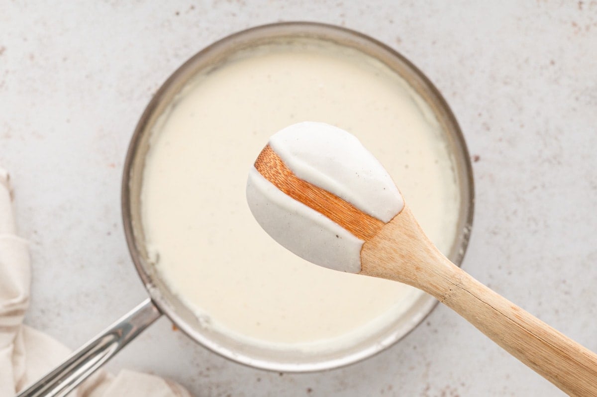 Alfredo sauce in a skillet, a wooden spoon with alfredo sauce on the back, with a scrape through the sauce to show how thick it is.